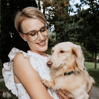 Junge Frau mit Brille, kurzem blonden Haar lächelt ihren goldenen langhaar Dackel an. Im Hintergrund ist Wald und die Sonne scheint.